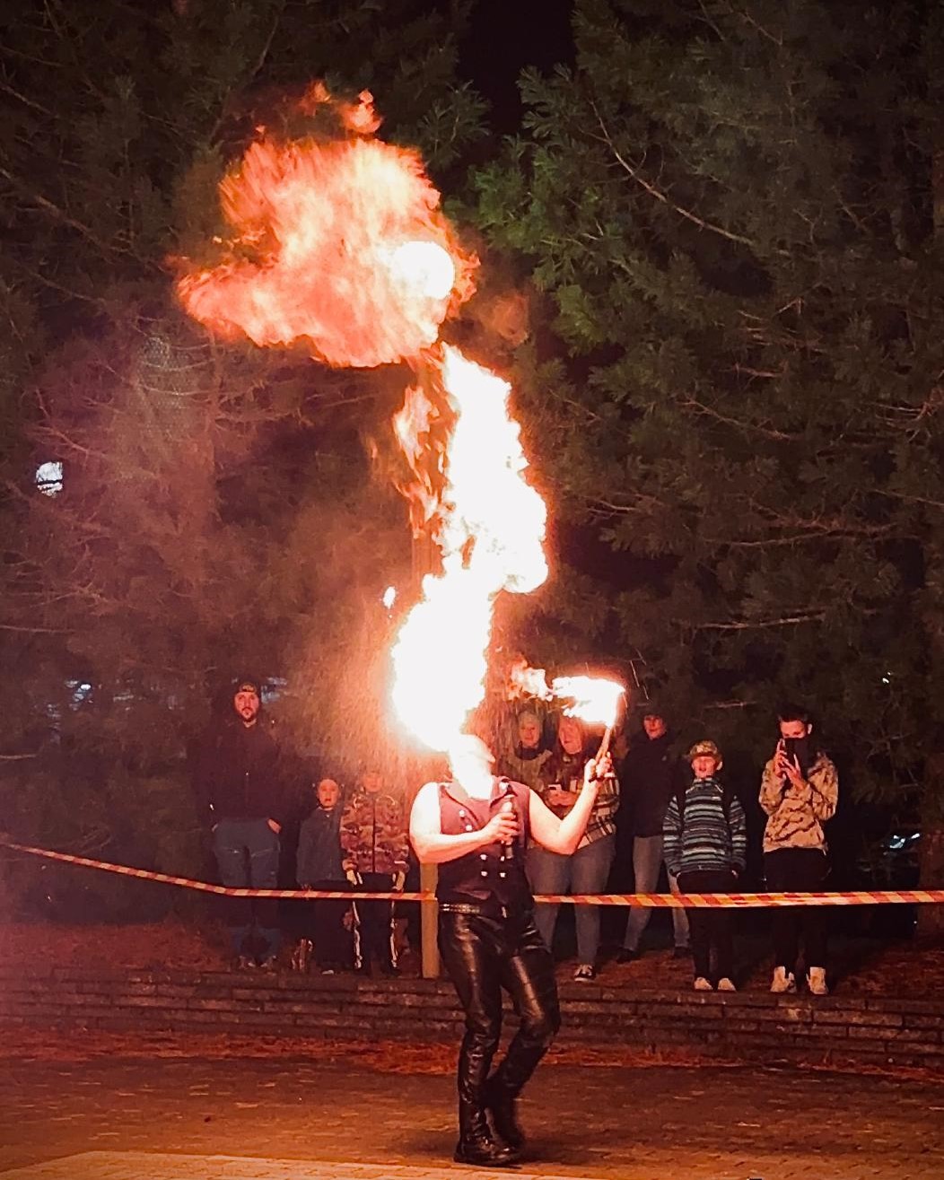 Ulkona pimeässä tuliesityksessä taiteilija puhaltaa ylöspäin ison liekin, nauhan takana näkyy yleisöä.